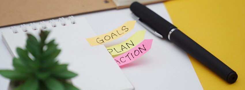 Scéna na stole obsahuje poznámkový blok a pero, ktoré zdôrazňujú riadenie času. Spod poznámkového bloku vykúkajú tri lepiace papieriky označené ako „CIELE“, „PLÁN“ a „AKCIA“. V popredí vľavo malá zelená rastlina dodáva tomuto organizovanému pracovnému priestoru nádych prírody.