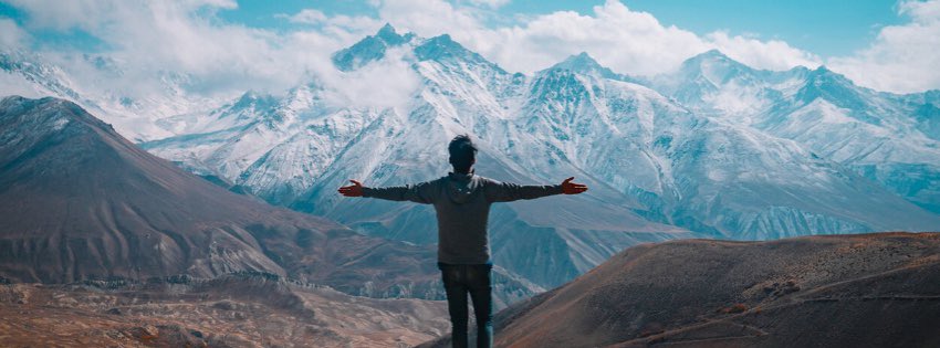 Osoba stojí s roztiahnutými rukami na hore a objíma panoramatický výhľad na zasnežené vrcholy pod polojasnou oblohou. Členitý terén v popredí kontrastuje s majestátnymi vzdialenými horami – inšpiratívna scéna pozývajúca na „vezmite si dovolenku.