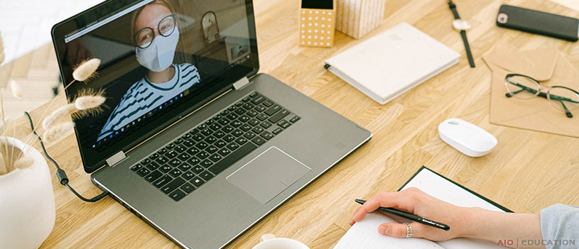 Osoba s okuliarmi a maskou telefonuje cez laptop a objíma návrat do práce po karanténe. Ruka držiaca pero píše do poznámkového bloku. Na drevenom stole nájdete aj myš, telefón a dekoratívne predmety.