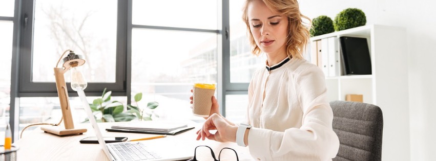Žena, dokonalá asistentka firmy, sedí za stolom vo svetlej kancelárii, kontroluje si náramkové hodinky a drží šálku kávy. Pred notebookom s okuliarmi a lampou nablízku efektívne zvláda úlohy. Poličky a okno tvoria pozadie jej pracovného priestoru.