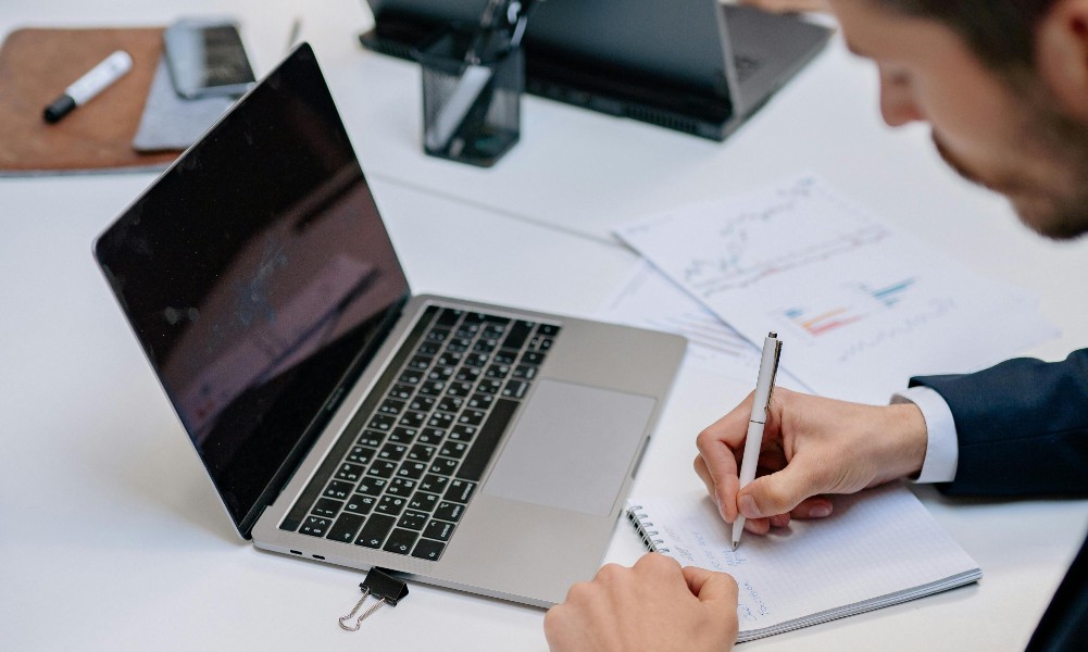 Pri stole plnom papierov, grafov a tabuliek človek píše do poznámkového bloku blízko otvoreného notebooku. Medzi nástrojmi produktivity, akými sú smartfón a pero, leží príprava na „hodnotiace rozhovory pre majstrov“, čo naznačuje prostredie zamerané na majstrovstvo a hodnotenie.