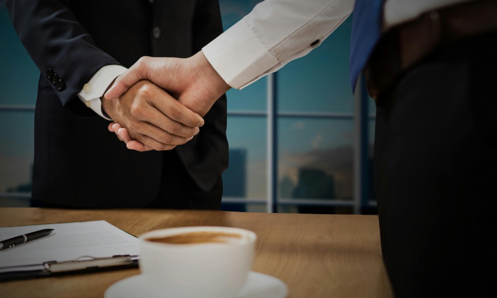 Dvaja ľudia v služobnom oblečení si podávajú ruky nad dreveným stolíkom so šálkou kávy a dokladom, ktorý naznačuje tréning pre pokročilých nákupcov. Okno odhaľuje výhľad na panorámu mesta, čo naznačuje, že prostredie je kancelária, kde sa uzatvárajú profesionálne dohody.