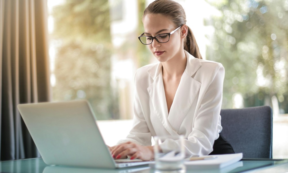 Osoba v okuliaroch a bielej blúzke, ktorá stelesňuje výkonnosť šikovnej asistentky, sedí za skleneným stolíkom a píše na notebooku. Zameraní na obrazovku si ich úloha vyžaduje silné komunikačné zručnosti. Pozadie je svetlé s rozmazanou zeleňou za oknom.