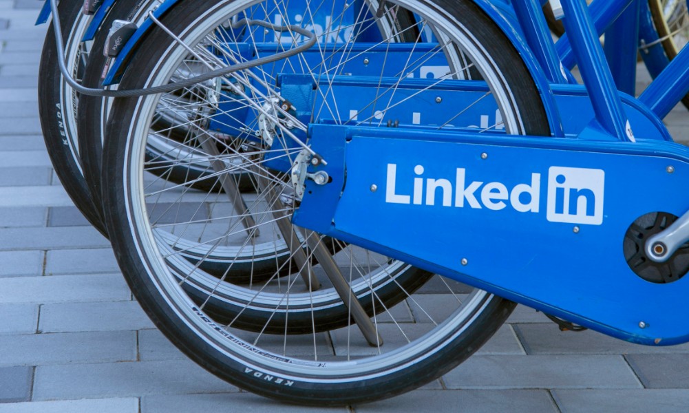 Detailný záber na niekoľko modrých bicyklov zaparkovaných v rade, pričom na každom je nápadne zobrazené logo LinkedIn na kryte zadného kolesa – dokonalý prvok značky pre obchodníkov. Na pozadí je vydláždená plocha so vzorom svetlých a tmavosivých dlaždíc.
