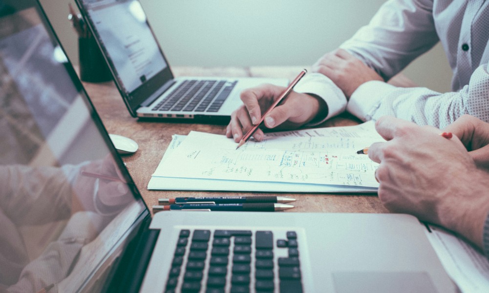 Dvaja ľudia pracujúci pri stole s dvomi otvorenými notebookmi sa zapájajú do diskusie o nákupe, keď jeden ukazuje ceruzkou na dokument, druhý pozorne počúva. Perá ležia vedľa dokumentu na drevenom stole, čo naznačuje, že táto kancelária alebo študijný priestor je zameraný na právo.