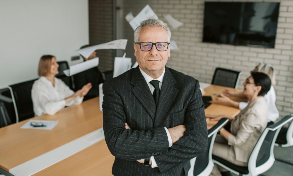 Starší muž v tmavom obleku a okuliaroch sebavedomo stojí so skríženými rukami na konci konferenčného stola a z jeho reči vyžaruje autorita. V pozadí sedia traja ľudia, ktorí komunikujú a animovane gestikulujú, pričom vo vzduchu sú viditeľné listy papiera.