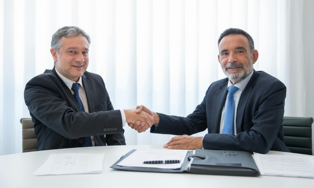 Dvaja muži v oblekoch si podávajú ruky cez stôl v kancelárskom prostredí, stelesňujúce úspešné vyjednávanie pre nákupcov. Dokumenty a priečinok sú úhľadne usporiadané na stole, zatiaľ čo priesvitný záves ponúka svetlé pozadie. Ich úsmevy odrážajú pozitívny výsledok ich rokovaní.