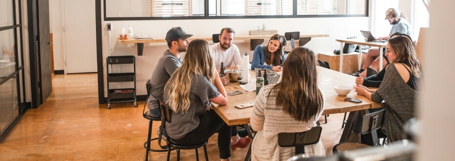 Okolo dreveného stola v modernom kancelárskom prostredí Centra sedí skupina ľudí a živo diskutuje. Niektorí majú otvorené notebooky na hodnotiace úlohy a na stole sú nápoje. Medzitým sa osoba v pozadí sústredí na svoj laptop pri samostatnom stole.