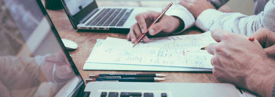 Dvaja ľudia spolupracujúci pri drevenom stole s otvorenými notebookmi, diskutujúci o tvorbe, ako jeden ukazuje na papier plný diagramov a hodnotiacich hárkov, zatiaľ čo druhý drží pero. Na stole vidno aj niekoľko ceruziek a zatvorený zápisník.