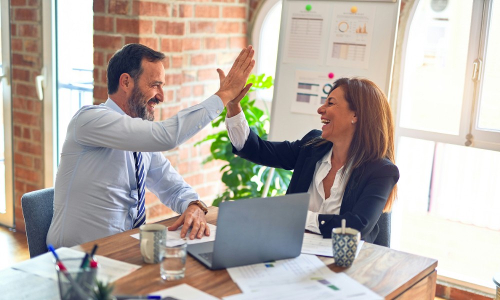 Muž a žena sediaci za stolom s notebookom a papiermi, stelesňujúci návyky na ľudí, sa usmievajú, keď si dávajú jeden druhého. Scéna sa odohráva na pozadí tehlovej steny, veľkého okna a bujnej rastliny.