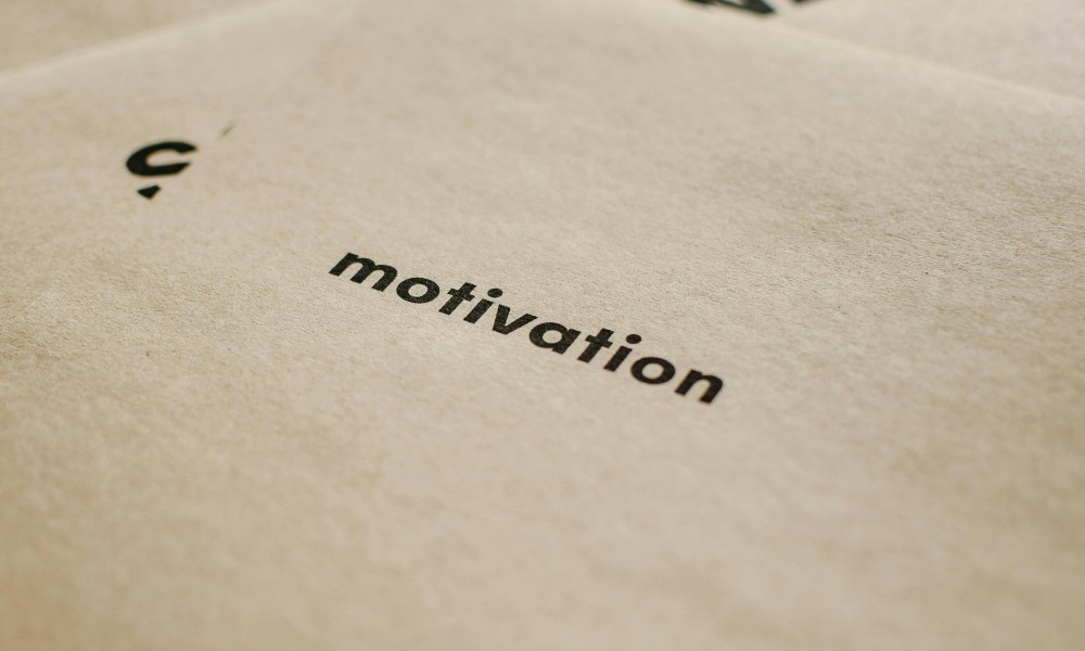 Snímka zachytáva detailný záber na kus papiera s elegantne vytlačeným slovom „motivácia“ v strede malými písmenami, čo odráža dynamiku motivovaných zamestnancov. Pozadie je jemné, svetlohnedé, dodáva scéne teplo.