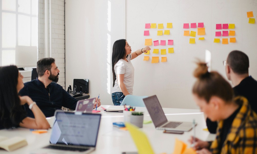 Žena stojí pri tabuli pokrytej farebnými lepiacimi papierikmi a ukazuje na jednu. Štyria kolegovia, súčasť dynamického manažérskeho mixu, sedia okolo stola s otvorenými notebookmi, perami a notebookmi a zúčastňujú sa tímového stretnutia v presvetlených, moderných kancelárskych priestoroch.