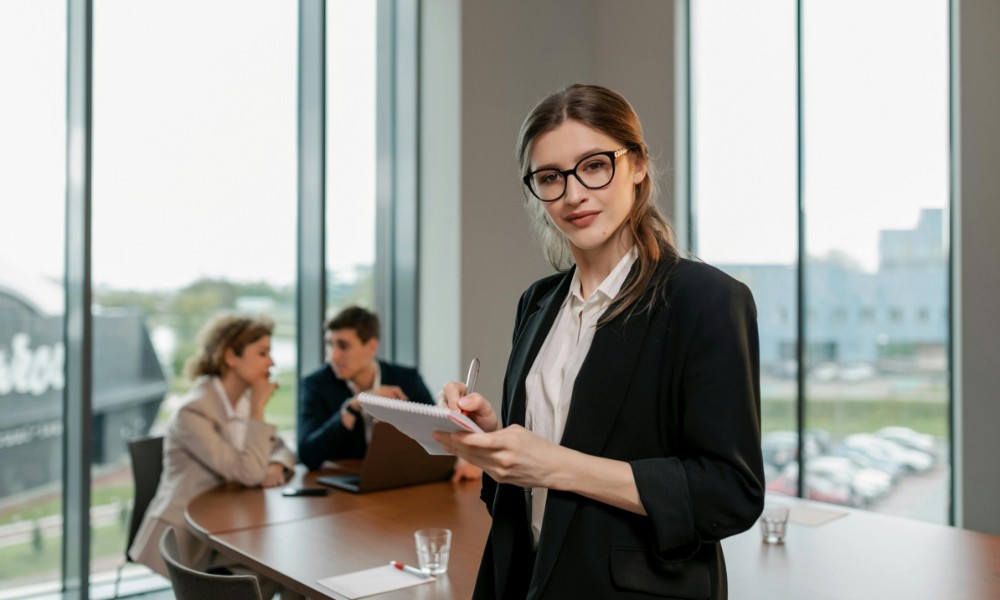 Žena v čiernom blejzri a okuliaroch sebavedomo stojí so zápisníkom a perom v ruke a je príkladom psychologického know-how vedúcich zamestnancov. V pozadí sedia dvaja ľudia za stolom pred veľkými oknami a rozprávajú sa.