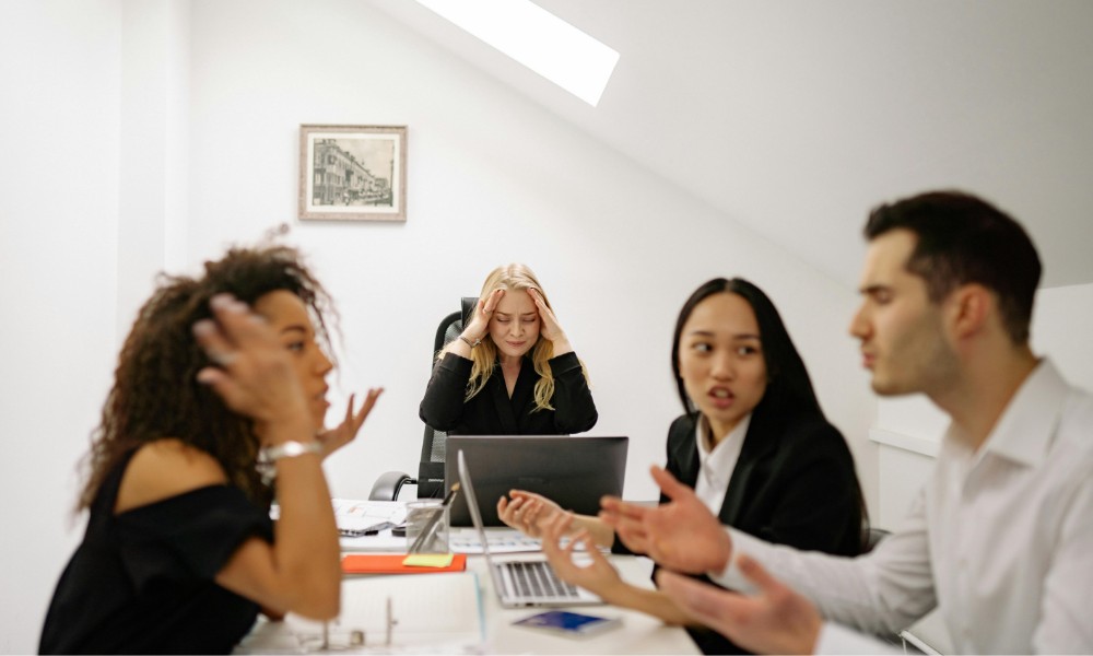 Skupina štyroch ľudí sedí okolo kancelárskeho stola. Jeden človek uprostred, zameraný na riešenie konfliktných situácií, sa chytí za hlavu a ostatní sa zapájajú do diskusie. Po stole sú rozhádzané notebooky a dokumenty.