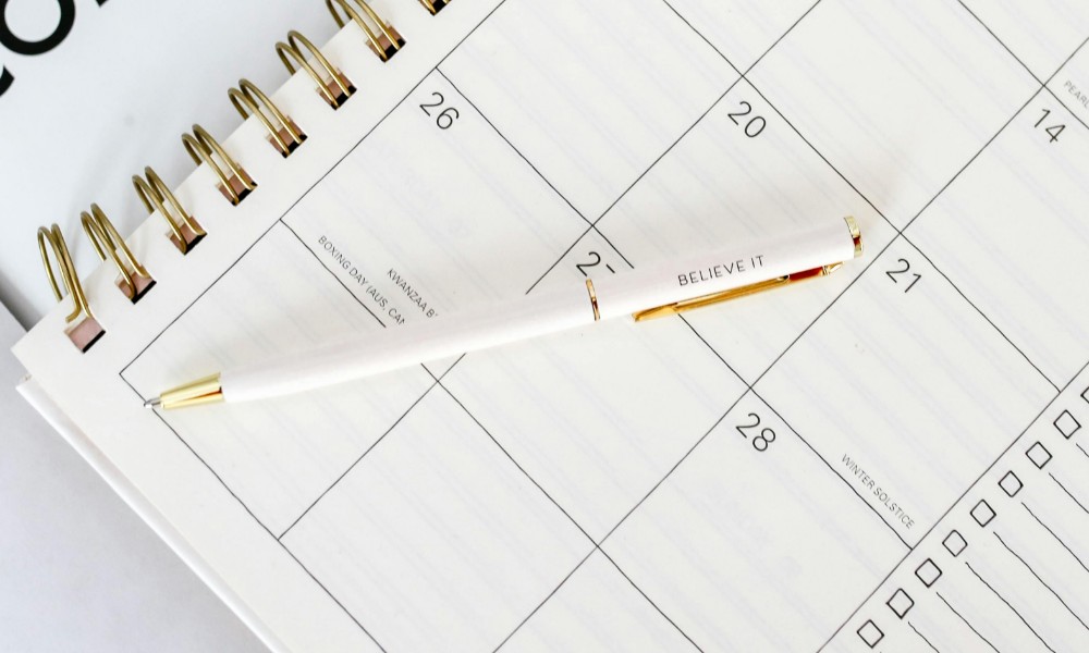 Bielo-zlaté pero s nápisom „BELIEVE IT“ leží uhlopriečne na špirálovitom plánovači, ktorý je otvorený na týždeň a zobrazuje dátumy vrátane 26. a 28. dňa. Tento čistý, minimalistický kalendár je základným nástrojom pre efektívne riadenie času.