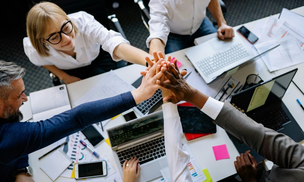 Rôznorodá skupina ľudí sedí okolo stola s notebookmi a dokumentmi a naťahuje ruky, aby položili ruky do stredu ako symbol tímovej spolupráce a spolupráce.