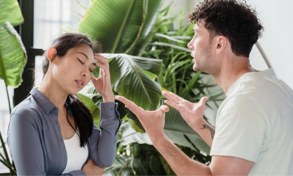 Muž animovane gestikulujúci pri rozhovore so ženou, ktorá vyzerá frustrovane s rukou na čele, demonštruje zvládanie konfliktu. Stoja pred veľkými zelenými rastlinami, žena v modrej košeli a muž vo svetlej.