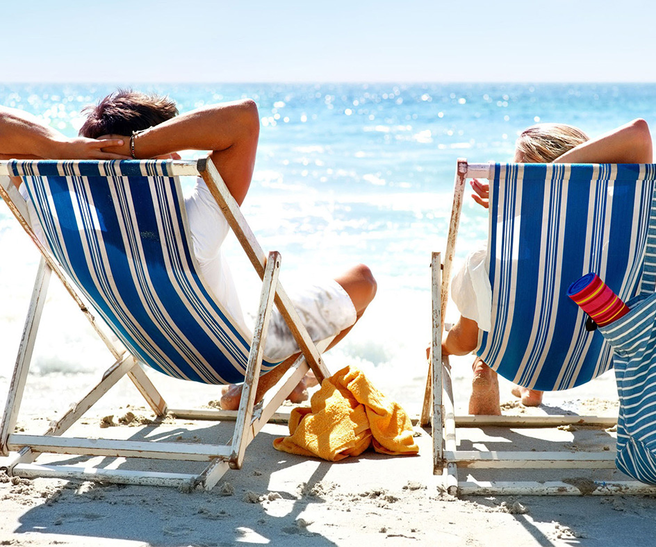 Dvaja ľudia relaxujú v pruhovaných plážových ležadlách na slnečnej piesočnatej pláži s výhľadom na oceán. Na piesku leží žltý uterák a v bočnom vrecku jednej stoličky je zastrčený nápoj. More je sýto modré pod jasnou oblohou – skutočne ideálna dovolenka.