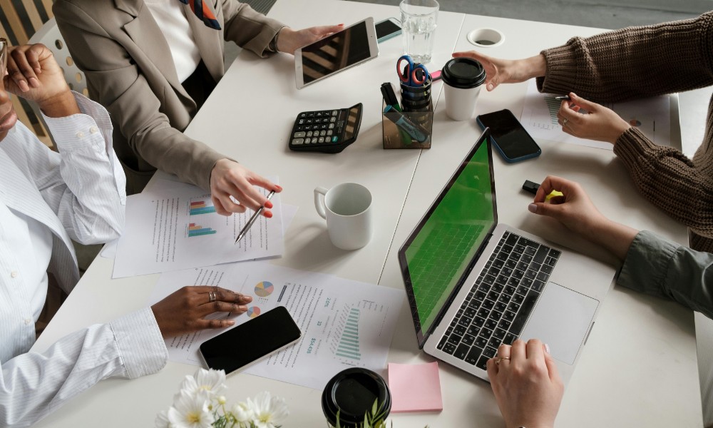 Skupina ľudí sediacich okolo stola sa zapojila do stretnutia zameraného na internú komunikáciu. Na stole sú rozhádzané papiere, grafy, kalkulačka, smartfóny a notebook. Niektorí gestikulujú a držia šálky kávy, zatiaľ čo jedna osoba píše na notebooku.