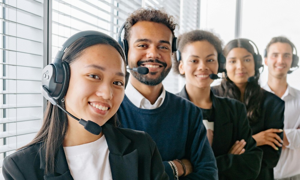 Rôznorodá skupina piatich ľudí v náhlavných súpravách a obchodnom oblečení stojí v rade s úsmevom. Stelesňujú efektívnu komunikáciu, čo naznačuje, že ide o call centrum alebo tím zákazníckych služieb. V pozadí sú vertikálne žalúzie.