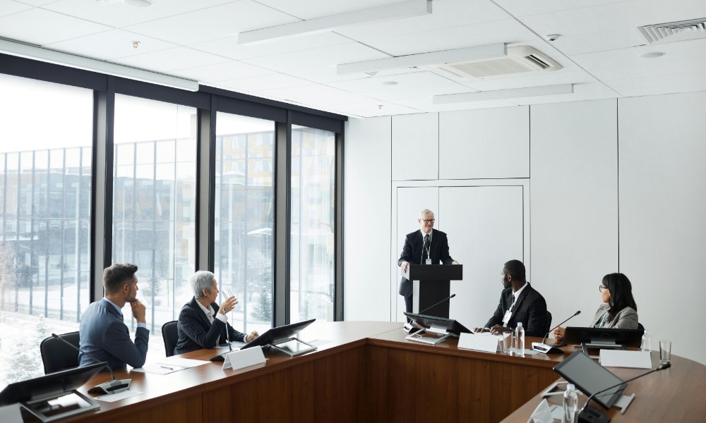 Skupina piatich ľudí v modernej konferenčnej miestnosti sa venuje kolektívnemu vyjednávaniu. Jedna osoba stojí na pódiu a hovorí, zatiaľ čo ostatní sedia pri veľkom drevenom stole a pozorne počúvajú. Veľké okná prepúšťajú prirodzené svetlo.