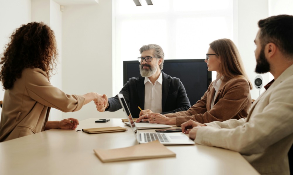 Štyria ľudia sú v zasadacej miestnosti, ktorá stelesňuje podstatu komunikácie ako základ úspechu. Žena a muž si cez stôl pevne potrasú rukou, zatiaľ čo ďalší dvaja sediaci kolegovia to pozorujú. Notebooky a dokumenty roztrúsené po okolí odrážajú ich spoločné úsilie.