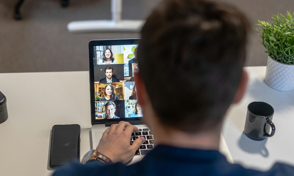 Osoba sediaca za stolom sa zúčastňuje videokonferencie na notebooku, kde predvádza efektívny komunikačný mix pre IT. Na obrazovke sa zobrazujú štyria ľudia v samostatných oknách videohovorov, zatiaľ čo stôl zdobí smartfón, čierny hrnček a zelená rastlina.