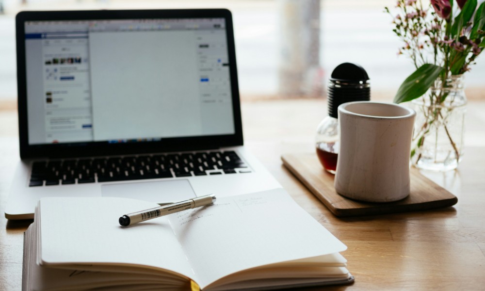 Notebook s webovou stránkou je otvorený na drevenom stole a stelesňuje efektívne firemné vzdelávanie. Vpredu vedľa neho leží otvorený zápisník s perom. Napravo je na drevenej tácke biely hrnček na kávu a malá váza s kvetmi.