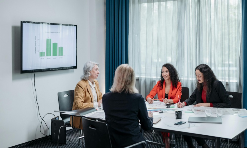 Štyria ľudia sedia okolo konferenčného stola v zasadacej miestnosti a diskutujú. Na nástennej obrazovke sa zobrazuje stĺpcový graf označený ako „akadémia pre personalistov“. Izba má veľké okná s modrými závesmi a na stole sú papiere a notebooky.