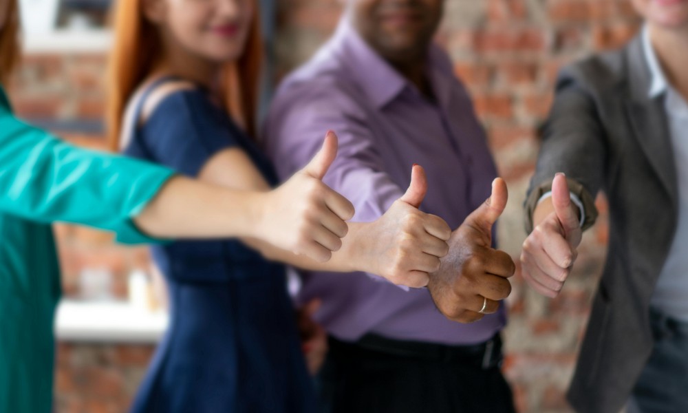 Skupina štyroch ľudí stojí vedľa seba a každý ukazuje palec hore smerom k fotoaparátu. Sú pred tehlovou stenou a stelesňujú osobný rozvoj. V centre pozornosti sú ich ruky, ktoré symbolizujú osobný rast prostredníctvom tohto pozitívneho a potvrdzujúceho gesta.