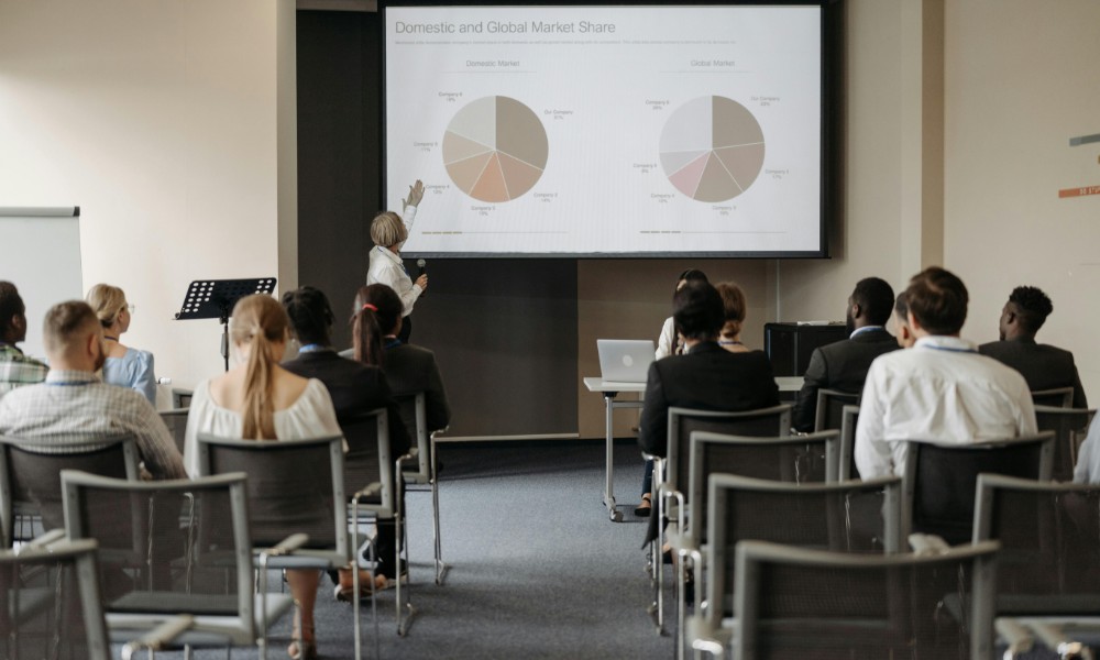 Moderátor s výnimočnými prezentačnými zručnosťami ukazuje na koláčové grafy na veľkej obrazovke v konferenčnej miestnosti. Účastníci sedia tvárou k obrazovke, niektorí si robia poznámky, zatiaľ čo iní pozorne počúvajú prezentáciu o domácom a globálnom podiele na trhu.