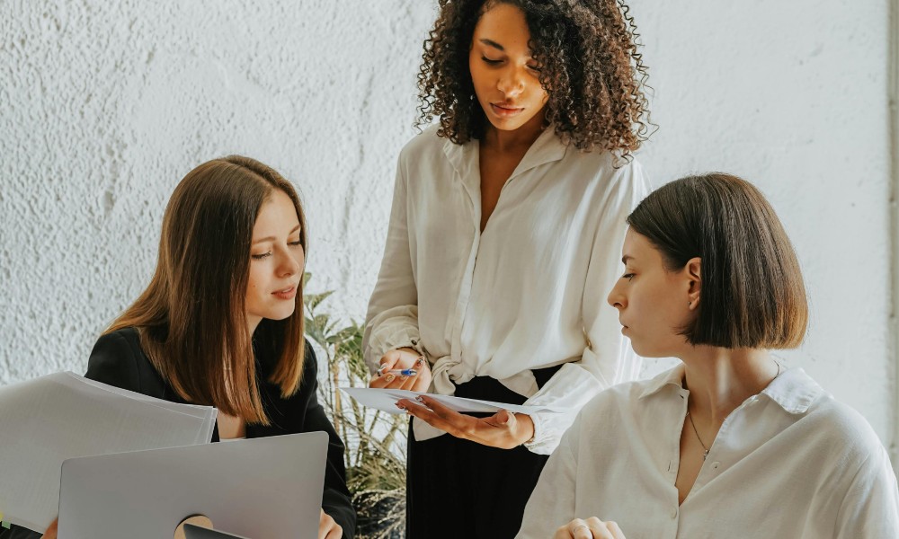 Tri ženy sa zapájajú do diskusie pri stole, využívajúc psychologické know-how pre personalistov. Jeden drží papiere, druhý sa pozerá na telefón, ktorý drží tretí, ktorý stojí. Sú vo svetlom kancelárskom prostredí s viditeľným notebookom a rastlinou.