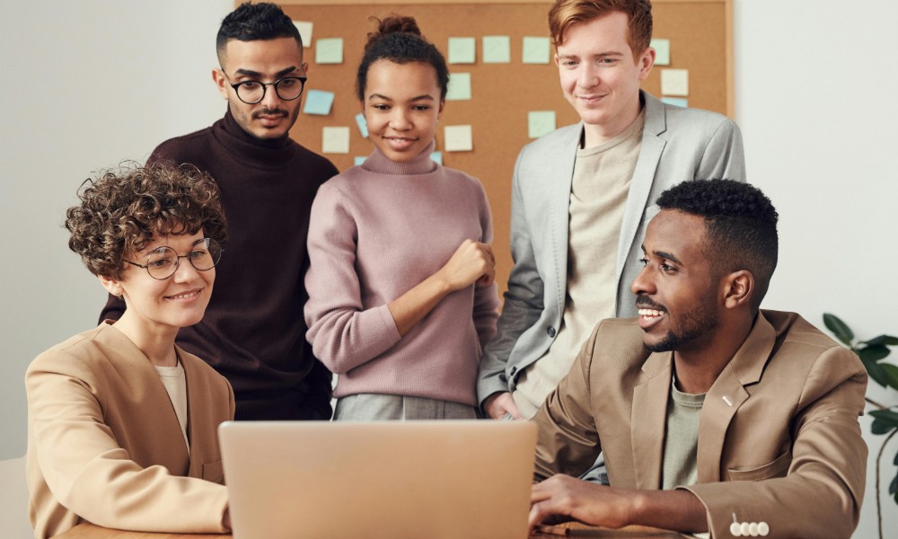 Rôznorodá skupina piatich ľudí stojí okolo notebooku, hlboko sústredená ako budovať firemnú kultúru. Nenútene oblečení sa zapájajú do živej diskusie s lepiacimi papierikmi na tabuli za nimi, ktoré naznačujú ich pracovný priestor alebo prostredie na stretnutia.