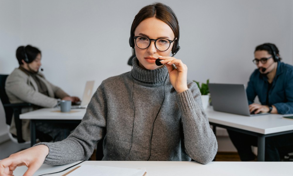Žena s okuliarmi a náhlavnou súpravou sedí za stolom v kancelárii a v sivom svetri sa venuje telefonickej a emailovej komunikácii. ona sa pozerá do kamery. Dvaja ďalší ľudia v náhlavných súpravách pracujú na notebookoch v pozadí.