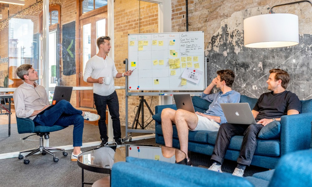 Štyria ľudia v modernom kancelárskom prostredí sa zameriavajú na zvládanie náročných situácií. Jeden stojí vedľa tabule pokrytej lepiacimi papierikmi a tabuľkami a pri rozprávaní gestikuluje. Ostatní traja sediaci s notebookmi na kolenách pozorne počúvajú. Izba má tehlové steny a veľké okná.