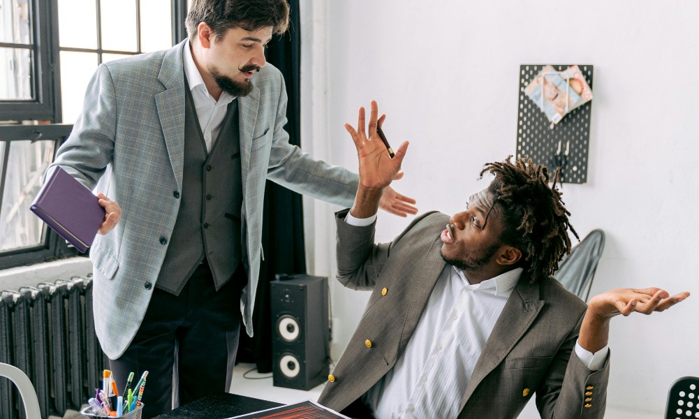 Dvaja muži v biznis oblečení sa pustili do búrlivej diskusie, možno strategicky zvládnutie náročných zákazníkov. Jeden stojí s knihou, expresívne gestikuluje, zatiaľ čo druhý sedí a krčí plecami so zdvihnutými rukami. V pozadí je viditeľná nástenka a vysoký reproduktor.