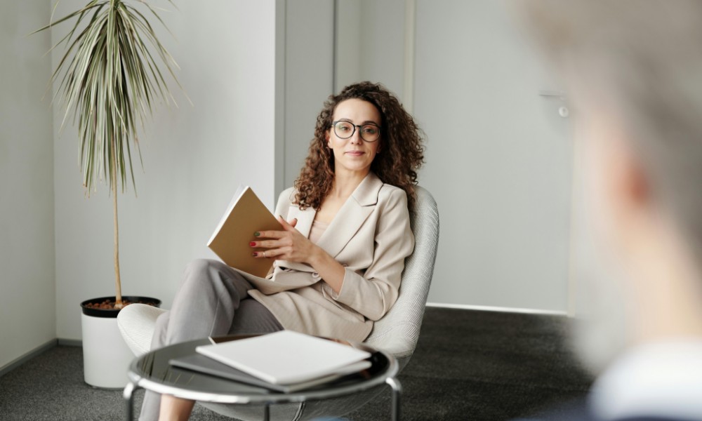 Žena s kučeravými vlasmi a okuliarmi sedí v kresle, drží notebook, stelesňuje podstatu manažéra ako kouč. Čelí niekomu čiastočne viditeľnému v popredí. Malá rastlina dodáva život do kancelárie, zatiaľ čo stôl zaprataný papiermi leží neďaleko.