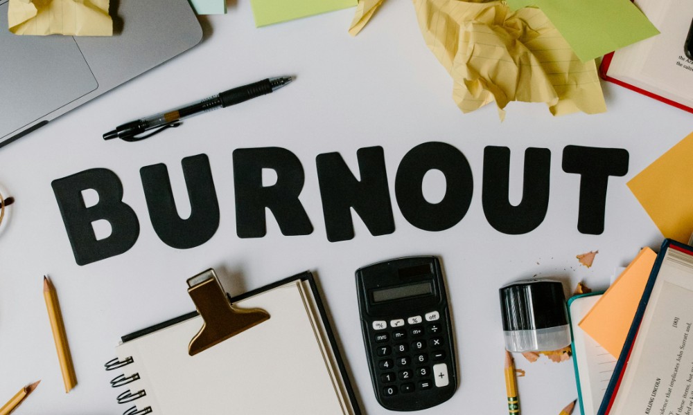 Zaprataný stôl s pokrčeným papierom, kalkulačkou, poznámkovým blokom, perami a pečiatkou obklopujú vystrihnuté písmená s nápisom „BURNOUT“, čo zdôrazňuje potrebu zvládania stresu v tomto ohromujúcom pracovnom alebo študijnom prostredí.